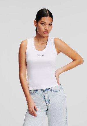 Top blanco sin mangas con textura acanalada y escote redondo, con un pequeño logotipo negro bordado cerca del pecho. Combinado con jeans de lavado claro.