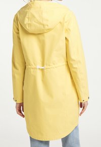 Femme portant un long imperméable jaune à capuche avec taille à cordon réglable, debout de dos portant un jean bleu clair.