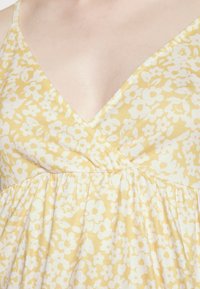 Vestido floral amarelo com padrões de flores brancas, design em forma de V e textura pregueada na cintura. Feito de um tecido suave e leve.