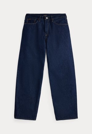 Jeune homme portant un maillot de rugby rayé violet et bleu marine, un jean bleu, des chaussures marron, un pull vert noué autour de la taille et des lunettes de soleil, debout devant un fond uni.