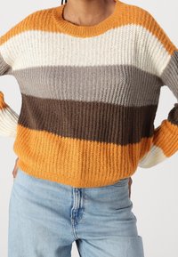 Person trägt einen gerippten Strickpullover mit horizontalen Streifen in Orange, Weiß, Grau und Braun, kombiniert mit hellblauen Denim-Jeans.