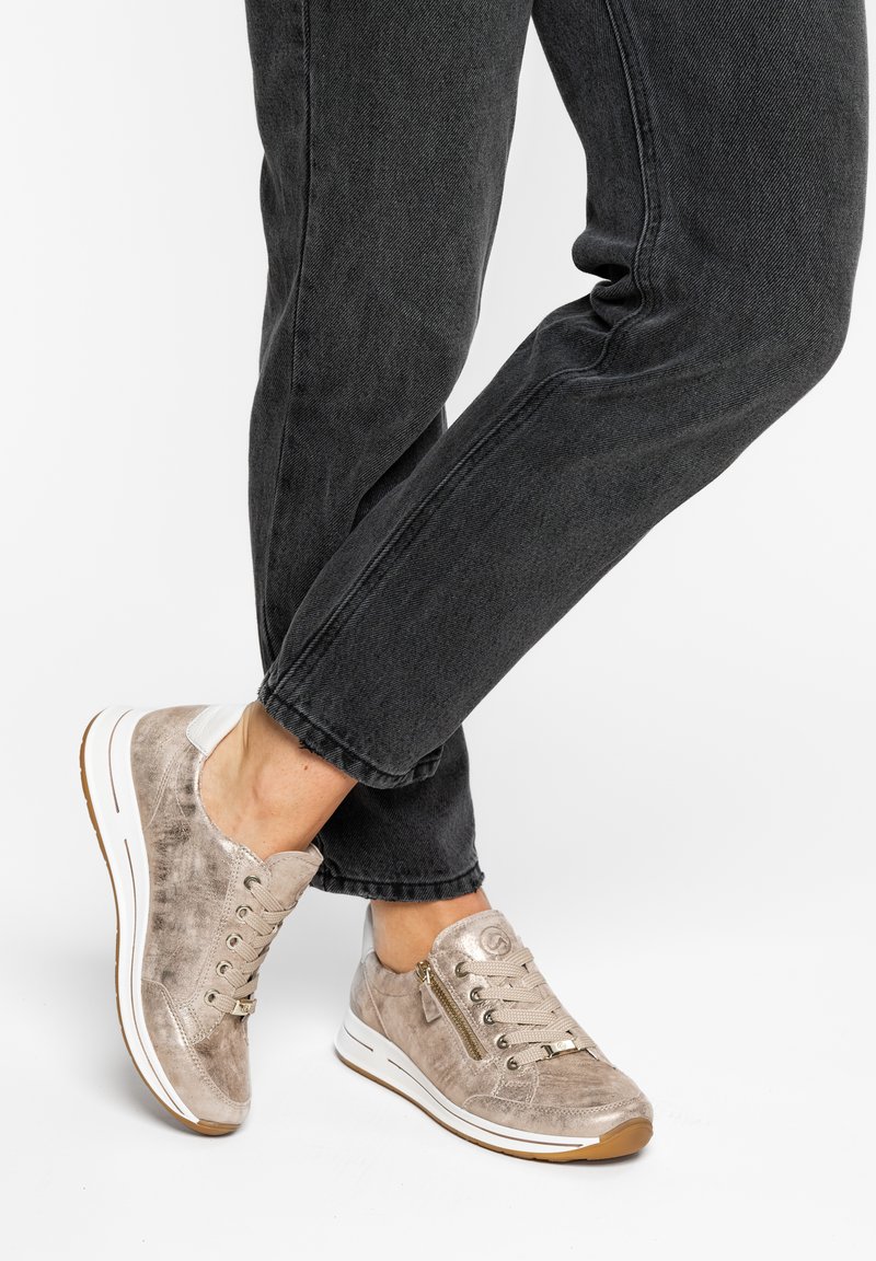 Persoon draagt beige suède sneakers met witte zolen en ritsen, gecombineerd met zwarte denim jeans tegen een witte achtergrond.