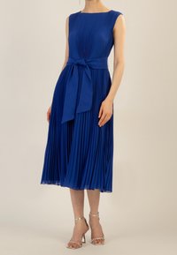 Blue pleated dress with a fitted bodice, round neckline, and waist tie. The flowing skirt reaches mid-calf with a smooth, sheer hem.