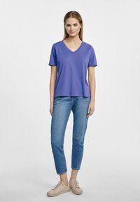 Fioletowa koszulka typu V-neck wykonana z miękkiej tkaniny, w zestawieniu z jasnoniebieskimi dżinsami i beżowymi sneakersami, posiada krótkie rękawy i luźny krój.