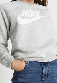 Šedá mikina s kulatým výstřihem a bílým logem Nike na hrudi; obsahuje žebrované manžety a měkkou, texturovanou tkaninu.