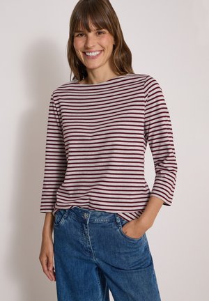 Femme souriante portant une chemise à manches longues rouge grenat et blanche à rayures et un jean bleu, une main dans la poche, sur un fond uni.