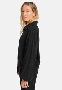 Zwarte corduroy pullover met een rits aan de voorkant, brede mouwen en een kraag. Heeft een ribstructuur en een geborduurd logo op de borst.