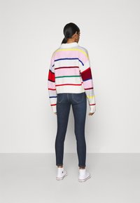 Pull en maille rayé multicolore avec des nuances de rose, rouge, vert et bleu, associé à un jean skinny foncé et des baskets blanches.