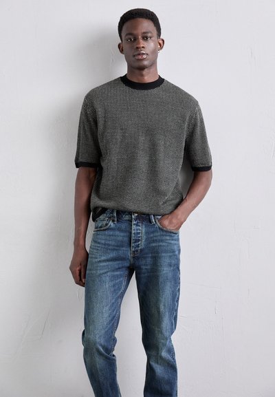 Pullover corto a maniche corte in bianco e nero con una trama, con scollatura rotonda, abbinato a jeans blu a gamba dritta, presentato su uno sfondo chiaro.