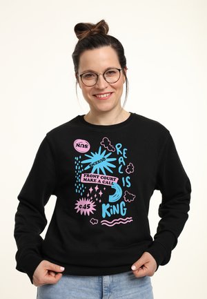 Smilende kvinde med briller iført en sort sweatshirt med blå og pink grafisk tekst: "Real er konge, front court tager beslutningen."
