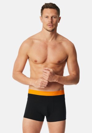 Skjorteløs mand iført sorte boxershorts med orange livrem, stående mod en ensartet lys baggrund, med hænderne foldet foran.