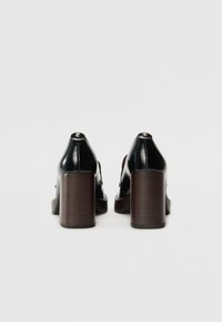 Chaussures à talons hauts en cuir noir avec un talon en bois, présentant un bout arrondi et des éléments décoratifs à l'arrière. Texture lisse.