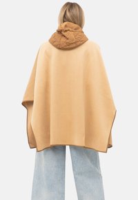 Vrouw die een beige cape met een bruin doorgestikt capuchon draagt, gecombineerd met lichtblauwe jeans met wijde pijpen, van de achterkant gefotografeerd.