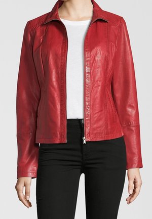 Mujer con chaqueta de cuero roja con cremallera sobre una camiseta blanca y pantalones negros, de pie frente a un fondo liso y claro.