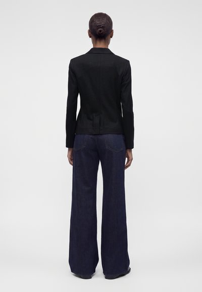 Femme avec les cheveux en chignon portant un blazer noir cintré et un jean large foncé, debout de dos sur un fond uni.