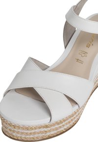 Sandale compensée blanche en cuir avec des lanières croisées, une plateforme tressée beige et dorée, et une finition lisse et texturée.