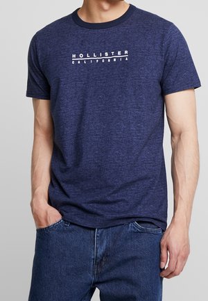 Mann trägt ein marineblaues Hollister California T-Shirt und blaue Jeans mit einer Hand in der Tasche vor einem schlichten grauen Hintergrund.