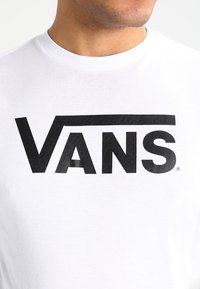 Valkoinen puuvillapaita, jossa on suuri musta "VANS" -logo erottuvalla alleviivauksella, pyöreä kaula-aukko ja sileä tekstuuri.