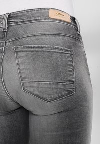 Los pantalones vaqueros de mezclilla gris tienen una cintura de altura media, bolsillos traseros y un desvanecimiento sutil. La etiqueta de la marca está cosida en la cinturilla.