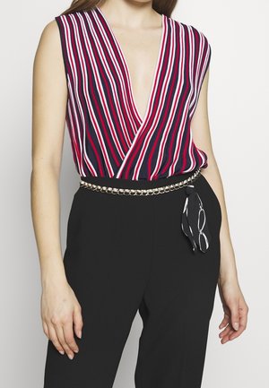 Ærmeløs top med V-hals med lodrette røde, hvide og marineblå striber, stoppet ned i højttaljede sorte bukser med et guldkæde bælte og tørklædeaccessory.
