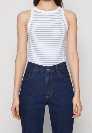 Kobieta ubrana w biały bezrękawnik z cienkimi niebieskimi poziomymi paskami oraz ciemnoniebieskie, wysokie dżinsy, stojąca na jednolitym tle.