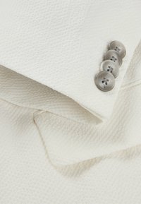 Tessuto blazer bianco texturizzato con revers e quattro bottoni grigi. Il tessuto mostra un motivo sottile e una finitura liscia.