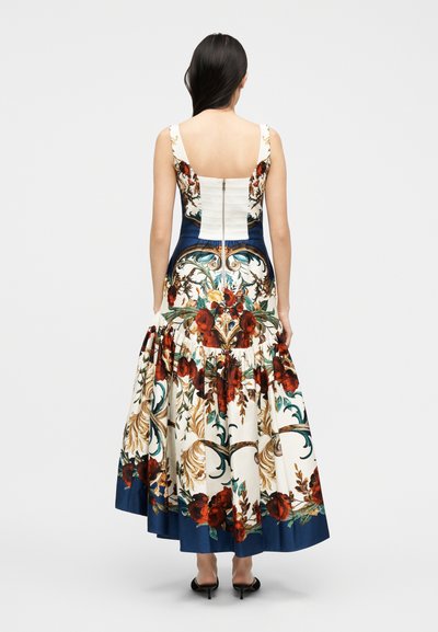 Femme portant une robe sans manches à motif floral avec un corsage ajusté et une jupe évasée, de dos, sur un fond uni.