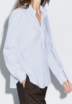 Kvinde iført en lyseblå skjorte med knapper og lange ærmer samt mørkebrune bukser, stående med den ene hånd i lommen mod en hvid baggrund.