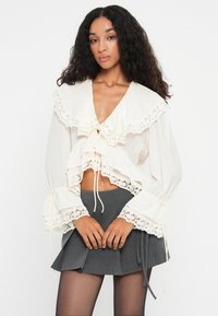 Witte blouse met gerimpelde kantafwerking, diepe V-hals en lange mouwen. Gepaard met een grijze rok, wat een gelaagd effect creëert.
