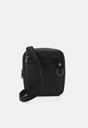 Czarna torebka Calvin Klein na ramię z kieszenią na zamek, przednim uchwytem na klucze i regulowanym, markowym paskiem na białym tle.