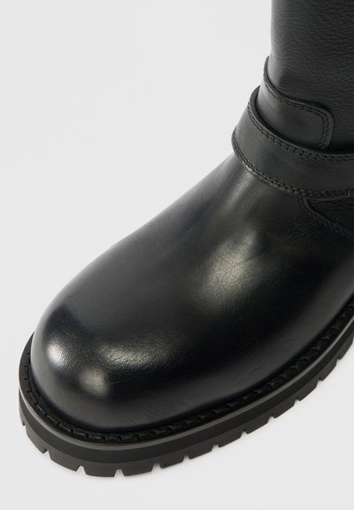 Μαύρη δερμάτινη μπότες με λεία επιφάνεια, στρογγυλή μύτη, και παχιά καουτσούκ σόλα με υφή. Λεπτομέρεια με λουρί στον αστράγαλο.