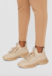 Chaussures de sport beige avec une tige en mesh texturé agrémentée d'accents métalliques et une semelle lisse et brillante. Portées avec des leggings beige clair ajustés.