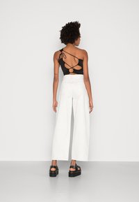 Top preto de um ombro com detalhe recortado, combinado com calças brancas de perna larga de cintura alta e sandálias de salto preto. Textura de tecido suave.