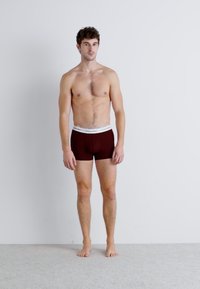 Burgundi színű boxer alsónadrág, kontrasztos fehér derékkal, puha anyagból készült. A modell mezítláb áll egy semleges háttér előtt.