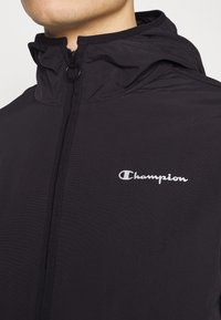 Black lightweight jacket with a zippered front, featuring a hood and a small white "Champion" logo on the chest. Smooth texture, minimal design.