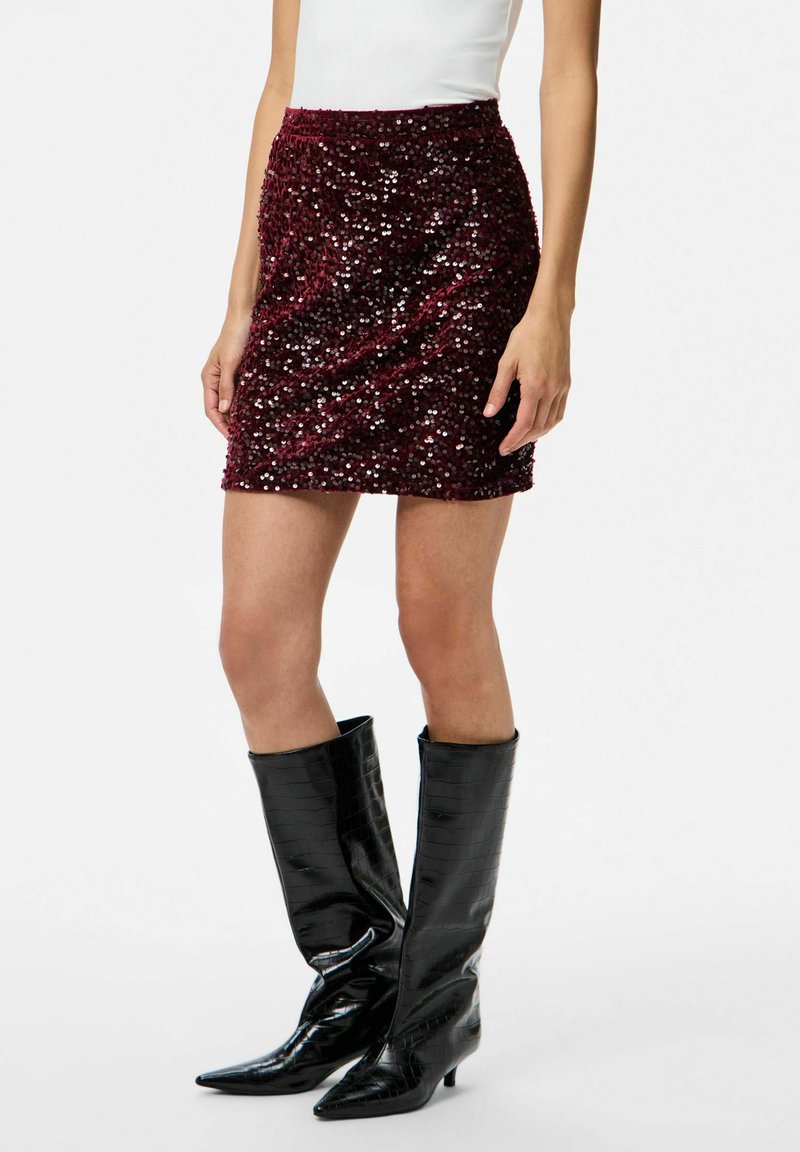 Mini falda de lentejuelas burdeos con cintura ajustada y superficie texturizada. Combinada con botas negras de charol por encima de la rodilla con punta afilada.