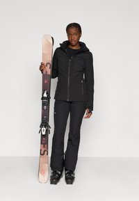 Équipement de ski comprenant une veste noire zippée avec des coutures obliques, un pantalon noir et des bottes de ski noires, associé à un ski marron clair avec des fixations noires.