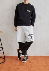 Osoba oblečená v černé mikině Moschino, bílých šortkách Moschino Milano navlečených přes černé pruhované kalhoty a černých teniskách s oranžovými akcenty.