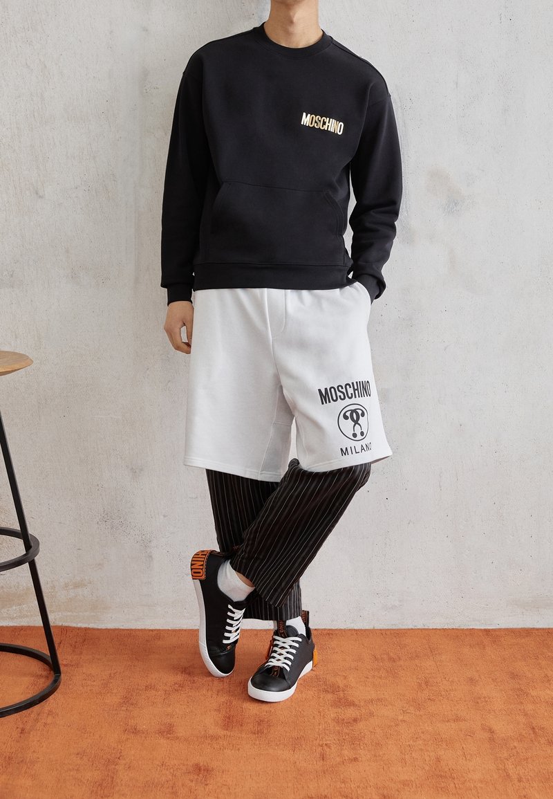 Osoba oblečená v černé mikině Moschino, bílých šortkách Moschino Milano navlečených přes černé pruhované kalhoty a černých teniskách s oranžovými akcenty.