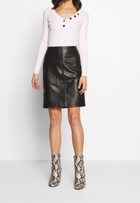 Mujer vestida con una blusa blanca de manga larga con botones, una falda de cuero negra hasta la rodilla y botas de tobillo con estampado de serpiente, de pie contra un fondo blanco.
