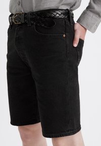 Pantalones cortos de mezclilla negra con corte recto, dobladillo deshilachado y diseño clásico de cinco bolsillos. Llevados con un cinturón de cuero negro trenzado.