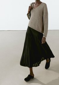 Pull en maille marron clair avec un col en V, associé à une jupe maxi plissée noire et des sabots noirs ornés d'accents métalliques.