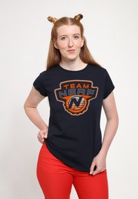 Tmavě modré tričko s grafikou "TEAM NERF" v oranžové a bílé barvě, krátké rukávy, uvolněný střih, doplněné červenými kalhotami. Hladká textura látky.