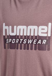Jasno-różowa bawełniana koszulka z dużym białym i ciemno-bordowym logo "hummel SPORTSWEAR" z teksturowanym wzorem pod spodem.