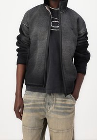 Osoba v tmavě šedé zipové bundě, černém tričku s grafikou, béžových vzorovaných kalhotách a perlou na krku, s rukama v kapsách.