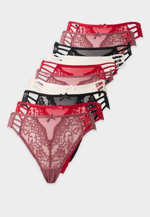 Femme debout pieds nus sur un fond blanc portant un soutien-gorge en dentelle noire assorti et une culotte avec des côtés à lanières.