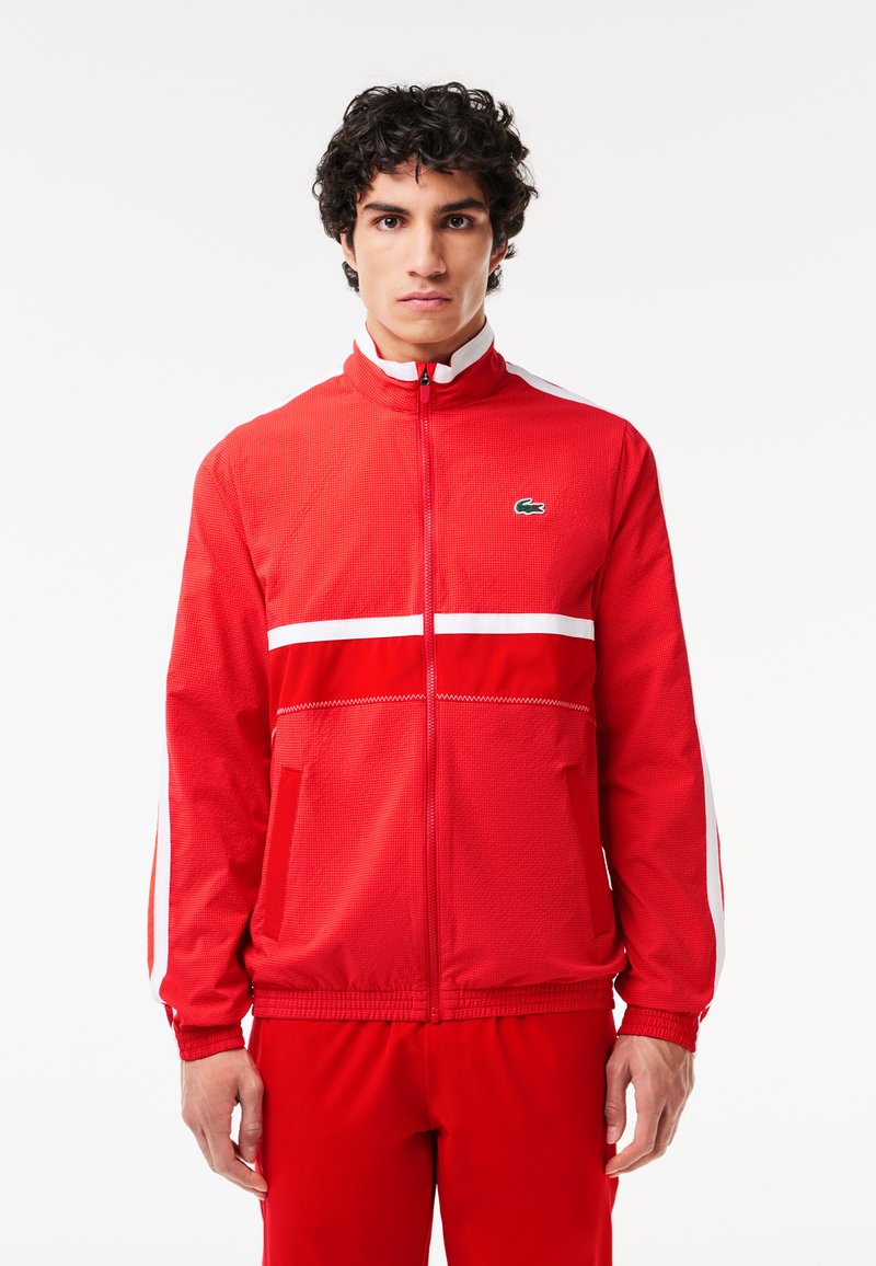 Veste zippée rouge en tissu texturé, avec des rayures blanches sur la poitrine et les manches, et un petit logo vert sur le côté gauche de la poitrine.