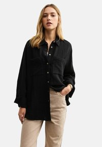 Zwarte blouse van textuurstof, met twee voorzakken, lange mouwen en een relaxte pasvorm. Gecombineerd met beige broek.