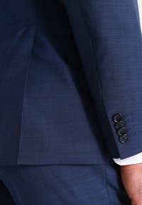 Abito blu navy con tessuto testurizzato; presenta un design a giacca monopetto e quattro bottoni sui polsini. Vestibilità sartoriale con linee nette.