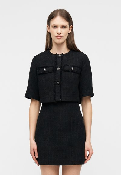 Νεαρή γυναίκα με ίσια καστανά μαλλιά φοράει μαύρο κοντό σακάκι με κοντά μανίκια και διακοσμητικά κουμπιά, σετ με φούστα ψηλόμεση που ταιριάζει.